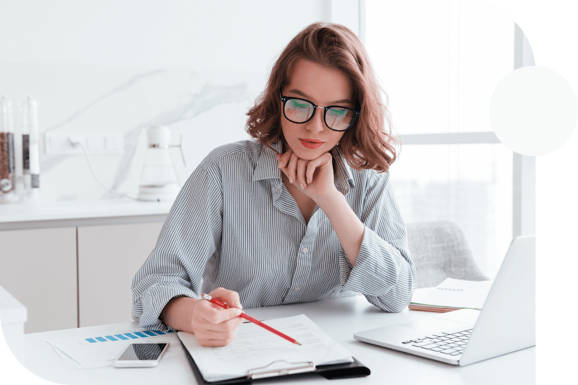 Person-wearing-glasses-sitting-at-a-desk-and-writing-something-on-a-piece-of-paper-with-their-laptop-open